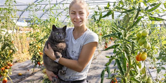 Porträt von Kati Fábián, die im Gewächshaus steht und einen grauen Kater auf dem Arm trägt.