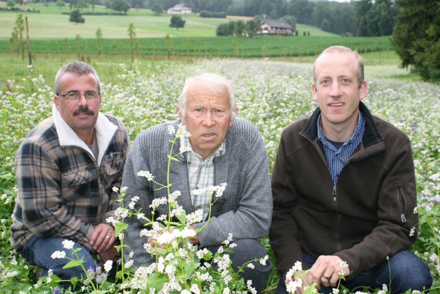 Hinten links die junge Nussbaum-Plantage, in der Mitte die Bienenweide, vorne drei Brückenbauer, von links: Nussbaum-Bauer Armin Lang, Imker Alois Leisbach und Simon Gisler von der Hochstamm Seetal AG. (Bild: zVg)