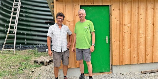 Rolf Mengelt und Andreas Braun (v. l.) stehen vor dem Pumpenhäuschen. Hinter ihnen ist ihr grünes Wasserreservoir zu sehen, das von Grundwasser gespeist wird. Von dort wird das Wasser in ihre Felder geleitet.