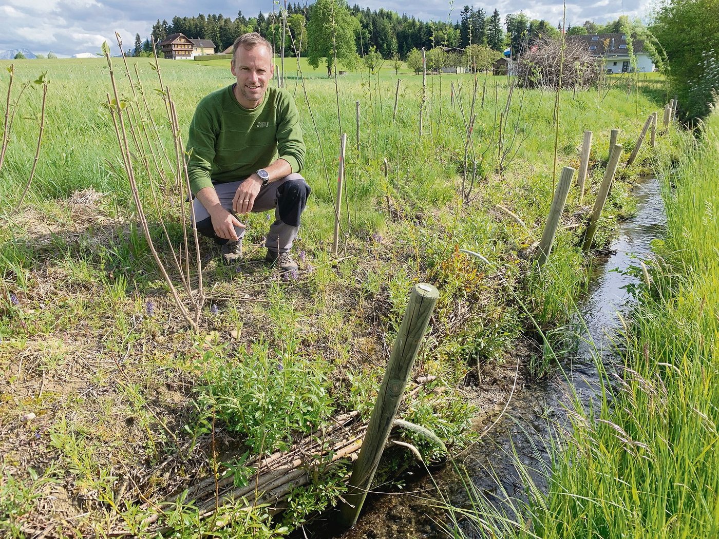 Landwirt Hubert Schürmann bei der neu angepflanzten Hecke entlang des Baches und Kleinstrukturen im Hintergrund. Vorne die Faschine als natürliche Bachverbauung. (Bild Josef Scherer)